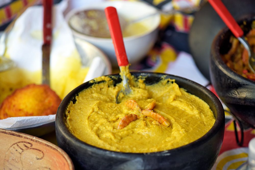 pote preto com vatapá, com uma colher dentro, em cima de uma mesa com vários outros pratos ao lado