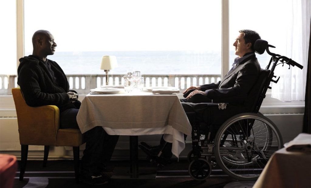 Os dois protagonistas do filme Os Intocáveis sentados à mesa do restaurante com vista para a cidade de Paris