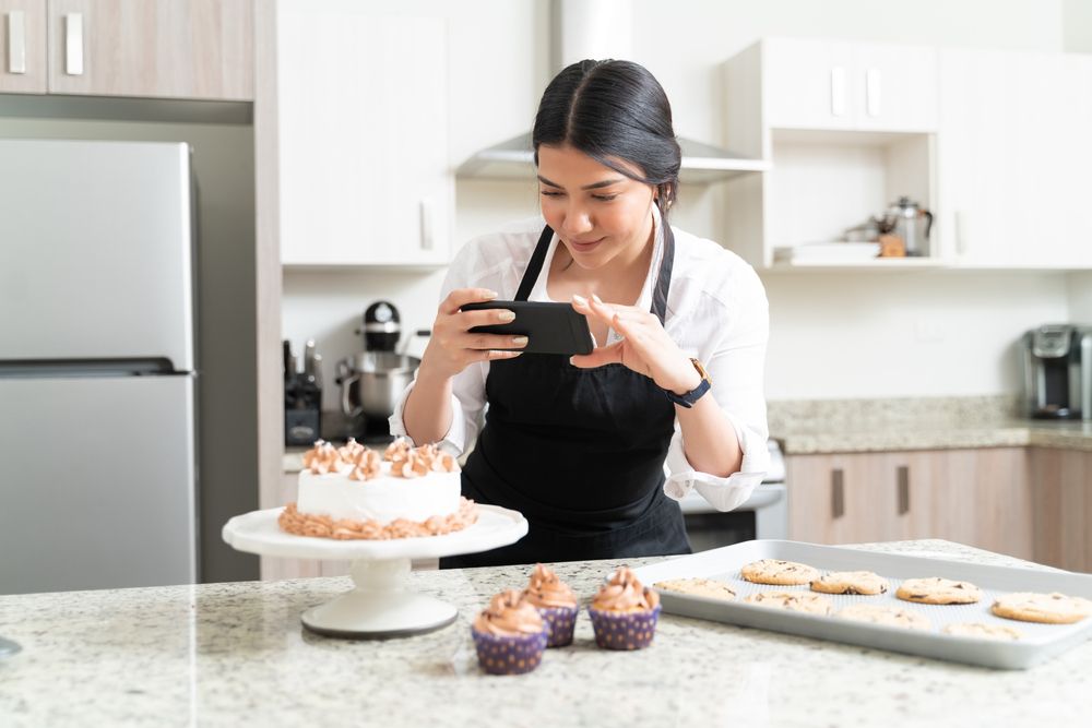 Mulher usando um avental preto tirando fotos de bolos confeitados com o celular com a cozinha de fundo