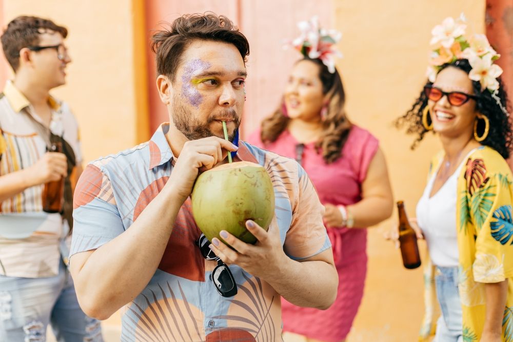 Homem com purpurina no rosto e camisa estampada tomando água de coco. Ao fundo, desfocados, um homem e duas mulheres tomam cerveja e usam adereços de carnaval.