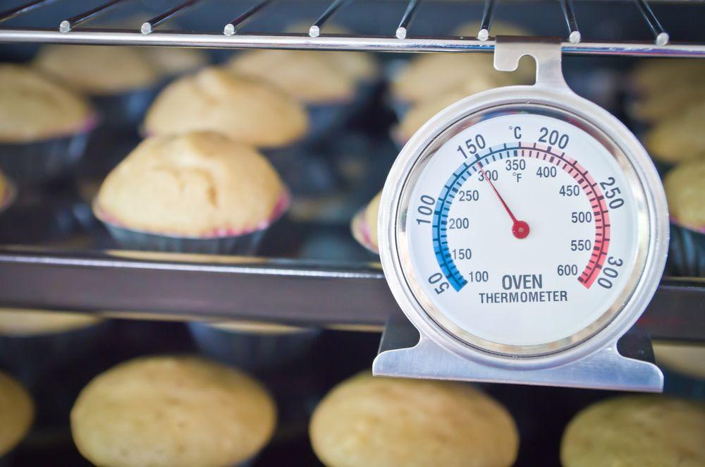 forno com um termometro pendurado na frente medindo a temperatura do forno e cupcakes atras