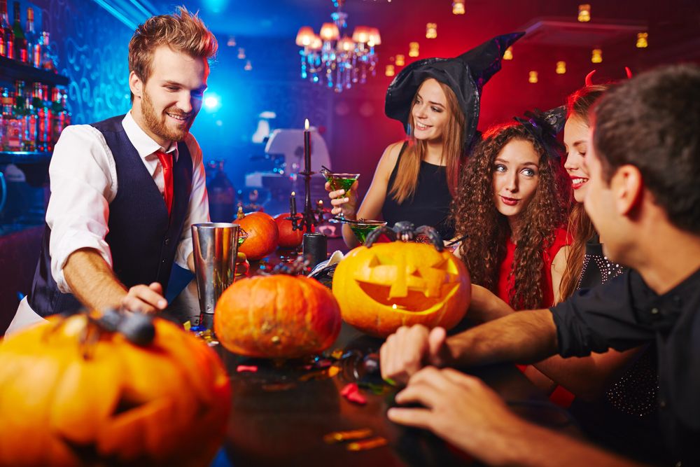 Quatro jovens com fantasias de bruxos em uma mesa de bar com abóboras de halloween com um barman servindo drinks