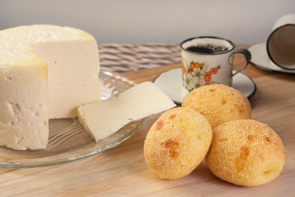 Foto de uma peça de queijo minas meia cura em um prato de vidro, ao lado de 3 pães de queijo e um xícara de café