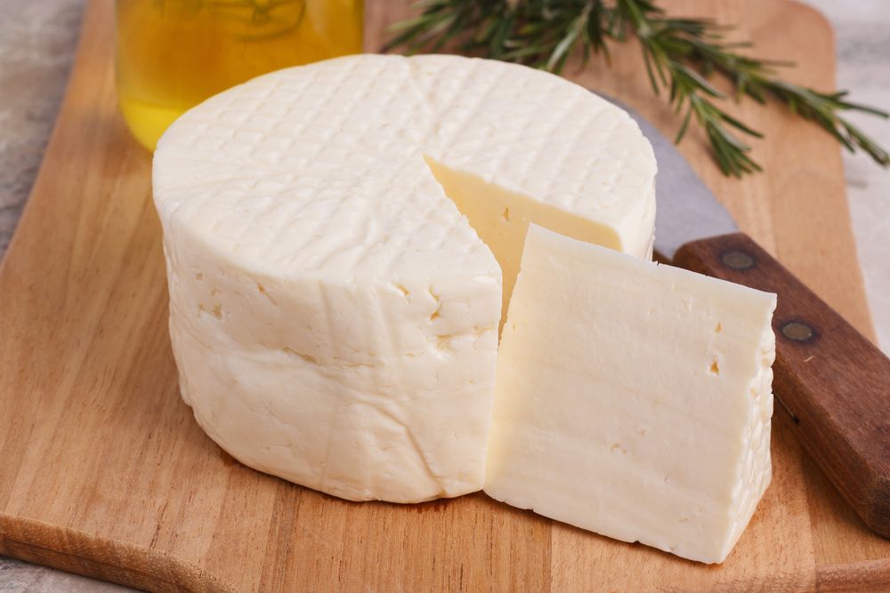 Imagem de um queijo minas meia cura com um fatia cortada, sobre uma mesa de madeira. Ao fundo, uma faca e um ramo de alecrim.