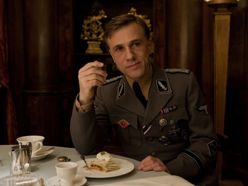 Coronel Hans Landa sentado à mesa com seu prato de apfelstrudel em cena no filme Bastardos Inglórios
