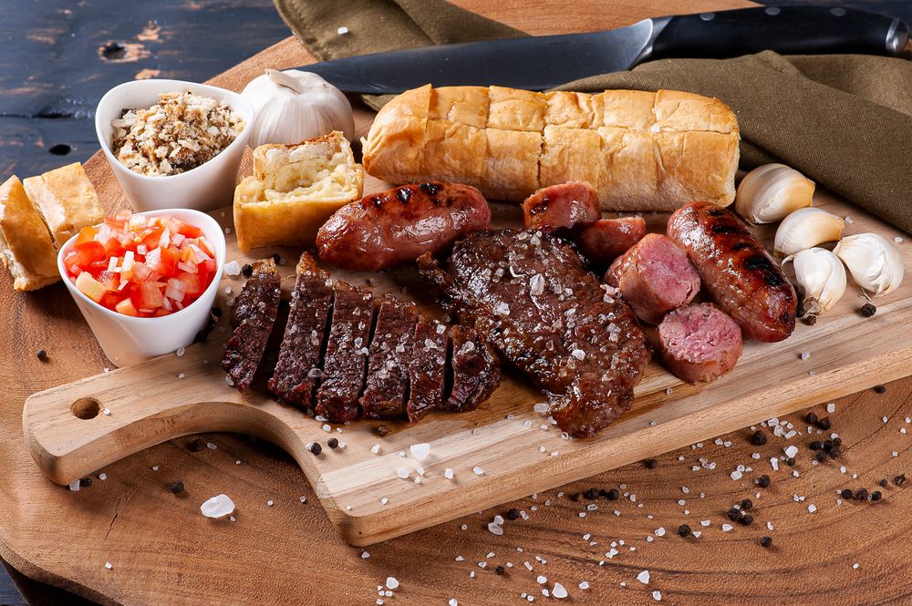 Uma imagem de Churrasco barato com várias carnes, pão e linguiça em uma tábua de madeira